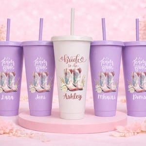 May include: Five personalized tumblers with lids and straws. Four are lavender, and one is white. Each features a cowboy boot design and the text "Team Bride" or "Bride to be". Names are printed on the tumblers.