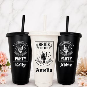 May include: Three personalized tumblers with lids and straws. Two black tumblers feature the text "TIL DEATH DO US" and "PARTY" with a skeleton hand graphic, and the names "Kelly" and "Abbie". The center white tumbler says "BRIDE OR DIE" and "Amelia".