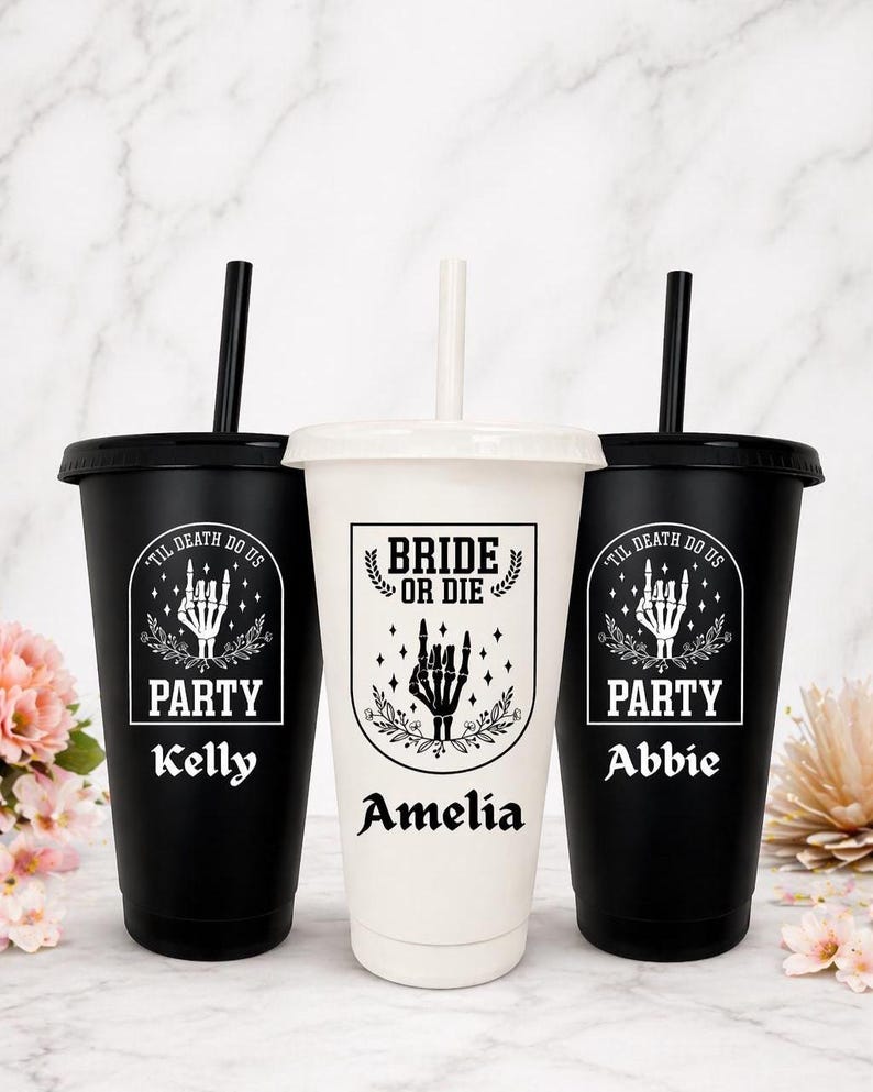 May include: Three personalized tumblers with lids and straws. Two black tumblers feature the text "TIL DEATH DO US" and "PARTY" with a skeleton hand graphic, and the names "Kelly" and "Abbie". The center white tumbler says "BRIDE OR DIE" and "Amelia".