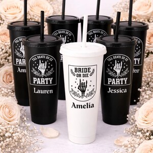 May include: A collection of black and white tumblers with lids and straws. The black tumblers have the text "TILL DEATH DO US PARTY" and names. The white tumbler has the text "BRIDE OR DIE" and a name. The tumblers are surrounded by flowers.