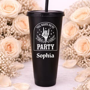 May include: Black tumbler with a lid and straw, featuring the text "TILL DEATH DO US PARTY Sophia" in white. The design includes a skeleton hand and floral accents. The background is decorated with roses and baby's breath.