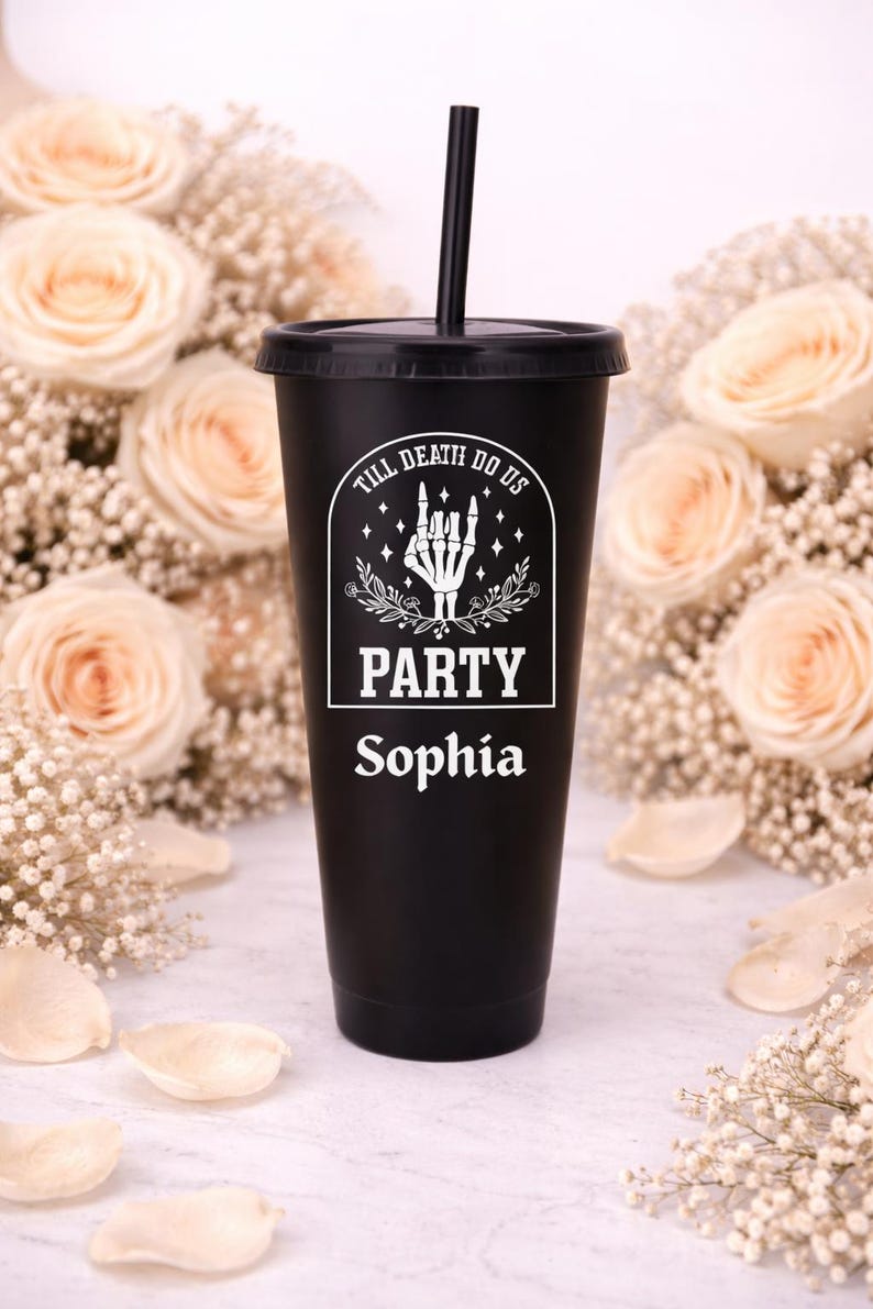 May include: Black tumbler with a lid and straw, featuring the text "TILL DEATH DO US PARTY Sophia" in white. The design includes a skeleton hand and floral accents. The background is decorated with roses and baby's breath.
