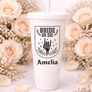 May include: White tumbler with a lid and straw, featuring the text "BRIDE OR DIE" above a skeleton hand graphic and the name "Amelia". The design is in black. The background includes roses and baby's breath.