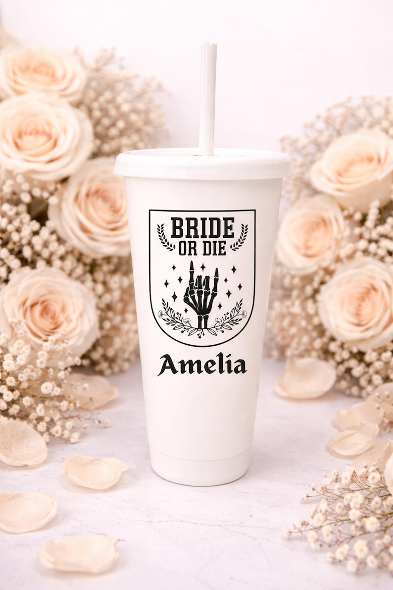 May include: White tumbler with a lid and straw, featuring the text "BRIDE OR DIE" above a skeleton hand graphic and the name "Amelia". The design is in black. The background includes roses and baby's breath.