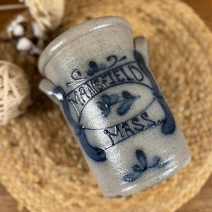 May include: A stoneware crock with a light grey base and blue lettering that reads "MANSFIELD MASS." The antique jar has two small handles and a decorative blue floral design. The crock is sitting on a woven mat.
