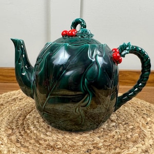 Puede incluir: Tetera de cerámica verde oscuro brillante, con forma de hoja de acebo, adornada con bayas rojas en la tapa y el asa. La tetera está colocada sobre una estera redonda de tejido.