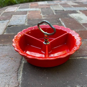 May include: Red ceramic divided serving dish with a metal handle. The dish has a scalloped edge and a decorative design.