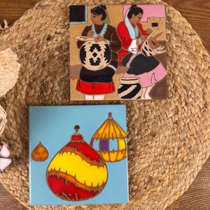 May include: Two decorative ceramic tiles. One tile features a scene of people working with baskets, in shades of brown, red, and black. The other tile has a light blue background with a red, yellow, and orange ornament design.