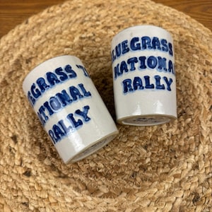 Puede incluir: Dos tazas de cerámica blanquecina con el texto "Bluegrass National Rally" en letras azules. Las tazas cilíndricas están sobre una estera redonda y tejida. Las tazas son probablemente vintage o hechas a mano.
