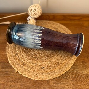 May include: A ceramic vase with a dark brown base and a blue and white top. The vase is placed on a round, woven mat. The vase has a glossy finish and a unique shape, suitable for home decor.