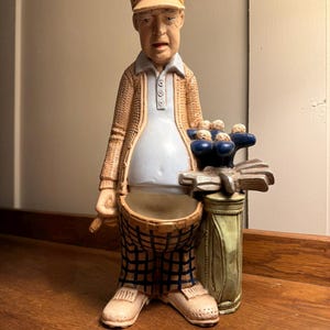 May include: A ceramic golf-themed figurine of a golfer wearing a tan cap and sweater, light blue shirt, and plaid pants. The figurine has a golf bag and golf balls. The figurine is on a wooden surface.