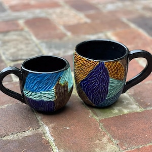 May include: Two ceramic mugs with abstract designs. The larger mug has a blue, purple, and orange design. The smaller mug has a blue, green, and brown design.