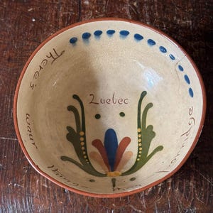 Puede incluir: Un pequeño cuenco de cerámica de color crema con un diseño floral azul, verde y naranja. El cuenco tiene un borde marrón y las palabras "There's a warm" y "fire in the" están escritas alrededor del borde. La palabra "Luebec" está escrita en el centro del cuenco.