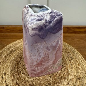 May include: A rectangular ceramic vase with a textured surface. The vase has a colour gradient, from deep purple at the top to pink and white at the base. It sits on a woven jute mat. The vase is approximately 20 cm tall.
