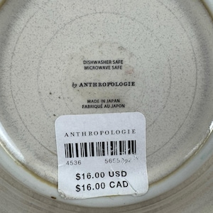 Puede incluir: Un plato de cer&aacute;mica con una superficie texturizada de color blanquecino. El plato tiene un borde ligeramente elevado y presenta el texto "DISHWASHER SAFE MICROWAVE SAFE by ANTHROPOLOGIE MADE IN JAPAN". Tambi&eacute;n es visible una etiqueta de precio.