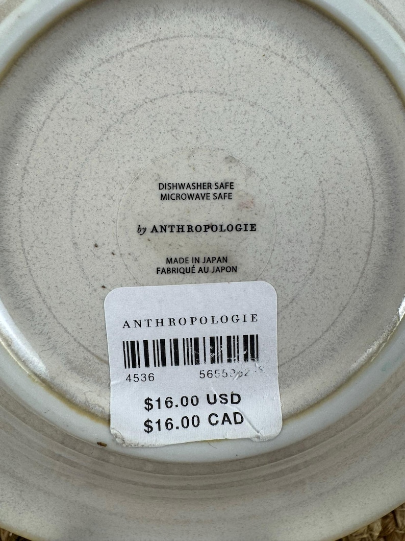 Puede incluir: Un plato de cer&aacute;mica con una superficie texturizada de color blanquecino. El plato tiene un borde ligeramente elevado y presenta el texto "DISHWASHER SAFE MICROWAVE SAFE by ANTHROPOLOGIE MADE IN JAPAN". Tambi&eacute;n es visible una etiqueta de precio.