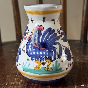 May include: A small white ceramic vase with a blue and yellow rooster design. The vase has a round body and a narrow neck.