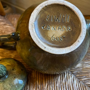 May include: A brown and green glazed ceramic teapot with a lid. The bottom of the teapot has the text "El Will Canada 605" printed on it.