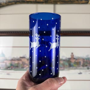 May include: A cobalt blue glass tumbler with white reindeer silhouettes and white polka dots. The cylindrical tumbler has a hand-painted appearance. The design suggests a festive, winter theme.