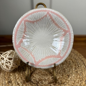 Puede incluir: Peque&ntilde;o plato redondo de cer&aacute;mica con interior blanco y dise&ntilde;o geom&eacute;trico rosa en el borde. Se exhibe sobre un soporte de metal dorado, mostrando su atractivo decorativo. Probablemente se utiliza para servir peque&ntilde;as porciones de comida o como adorno.