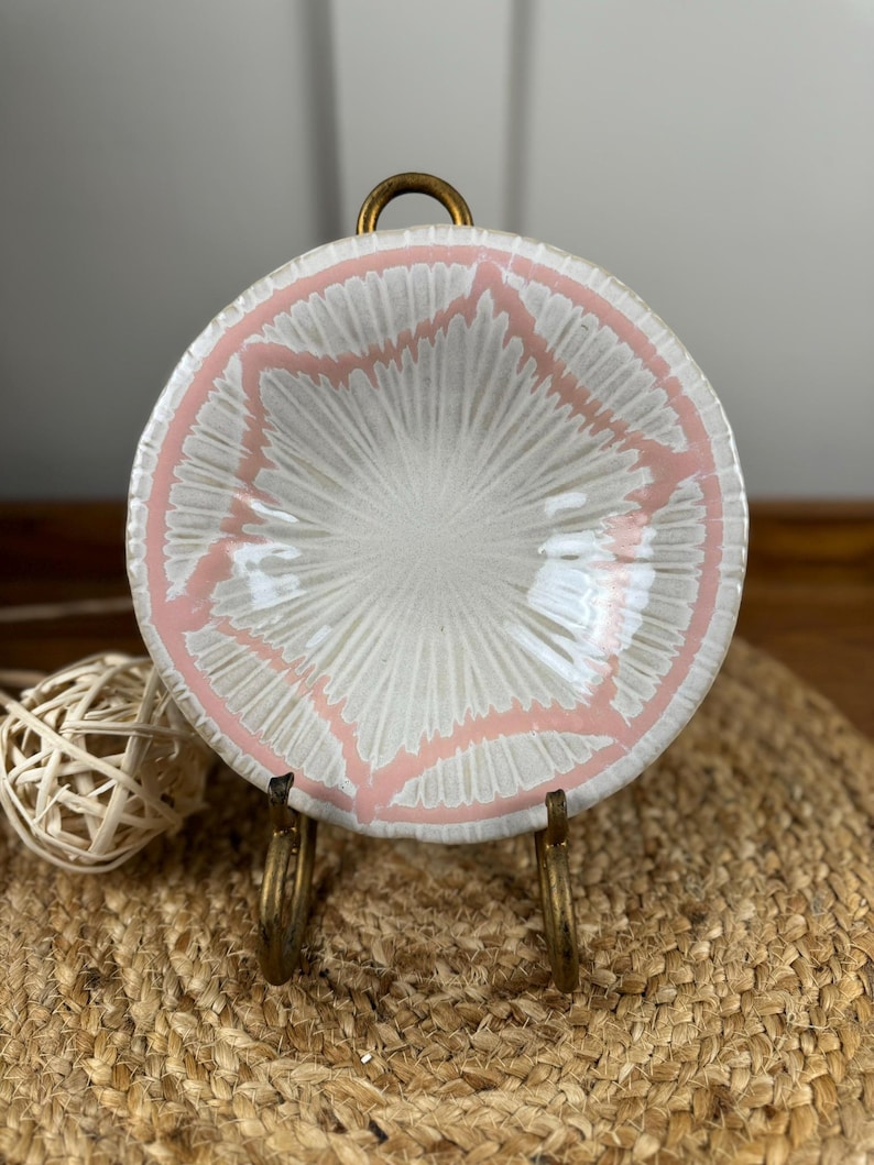 Puede incluir: Peque&ntilde;o plato redondo de cer&aacute;mica con interior blanco y dise&ntilde;o geom&eacute;trico rosa en el borde. Se exhibe sobre un soporte de metal dorado, mostrando su atractivo decorativo. Probablemente se utiliza para servir peque&ntilde;as porciones de comida o como adorno.