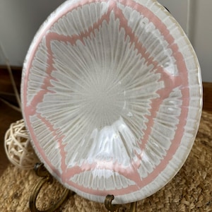 Puede incluir: Un plato de cer&aacute;mica blanco con un dise&ntilde;o de estrella rosa, exhibido en un soporte de metal dorado. El plato tiene una superficie texturizada y un borde ligeramente elevado. El soporte tiene un asa curva en la parte superior.