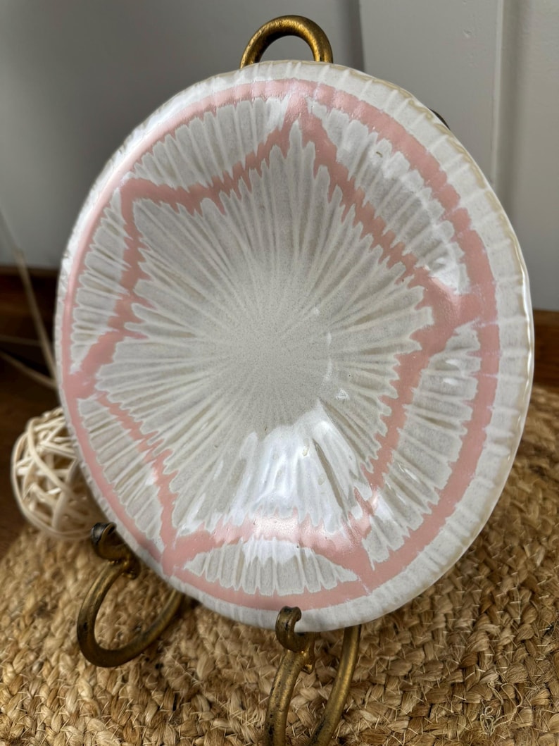 Puede incluir: Un plato de cer&aacute;mica blanco con un dise&ntilde;o de estrella rosa, exhibido en un soporte de metal dorado. El plato tiene una superficie texturizada y un borde ligeramente elevado. El soporte tiene un asa curva en la parte superior.