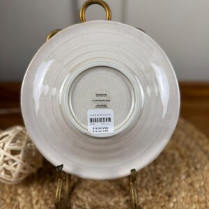 Puede incluir: Un plato Anthropologie beige claro, visto desde abajo, con una superficie texturizada. El plato se muestra en un soporte de metal dorado. El plato tiene un dise&ntilde;o circular con un borde ligeramente elevado.