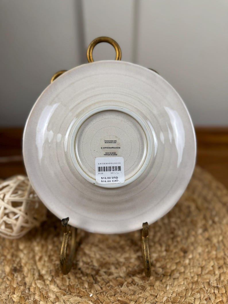 Puede incluir: Un plato Anthropologie beige claro, visto desde abajo, con una superficie texturizada. El plato se muestra en un soporte de metal dorado. El plato tiene un dise&ntilde;o circular con un borde ligeramente elevado.