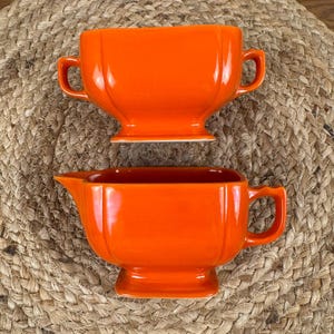 Puede incluir: Juego de servir de cerámica naranja de dos piezas. El juego incluye un cuenco con asa y una jarra para crema o salsa. Las piezas tienen un acabado brillante y están sobre una superficie tejida de tonos neutros.