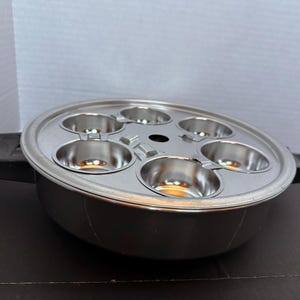 May include: A stainless steel egg poacher with six individual cups. The poacher has a round base and a lid with a central vent. Black handles are attached to the sides. The poacher is designed for cooking eggs.