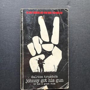 May include: A vintage paperback book with a black cover featuring a white peace sign and a silhouette of a soldier. The title "Johnny Got His Gun" is displayed in white text, with the author's name, Dalton Trumbo, above it. The text "The Most Shocking Anti-War Novel Ever Written!" is at the top.