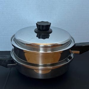 May include: Stainless steel cookware set with a lid and two black handles. The pot has a shiny, reflective surface and a black knob on the lid. The set is designed for cooking and food preparation.