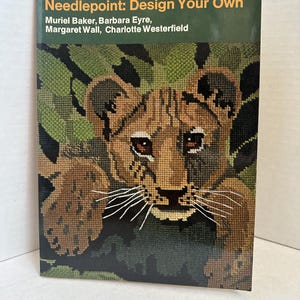 May include: A book titled "Needlepoint: Design Your Own" featuring a needlepoint design of a lion cub. The cover has a green background with the title and author names. The lion cub is brown and tan with white whiskers.