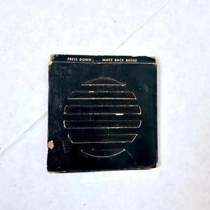 May include: A small, square, black card with the text "PRESS DOWN... MAKE BACK BULGE" at the top. The card features a series of horizontal lines forming a circular shape. The card has some wear and tear.
