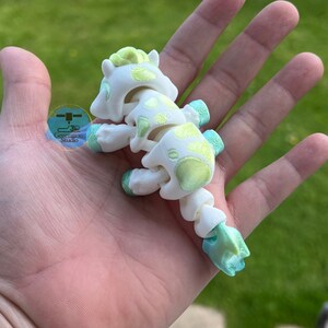 May include: A white articulated horse toy with green and yellow accents, held in a hand. The toy has a segmented body and a detailed design, with the text "Generagon Studio" visible in the background.