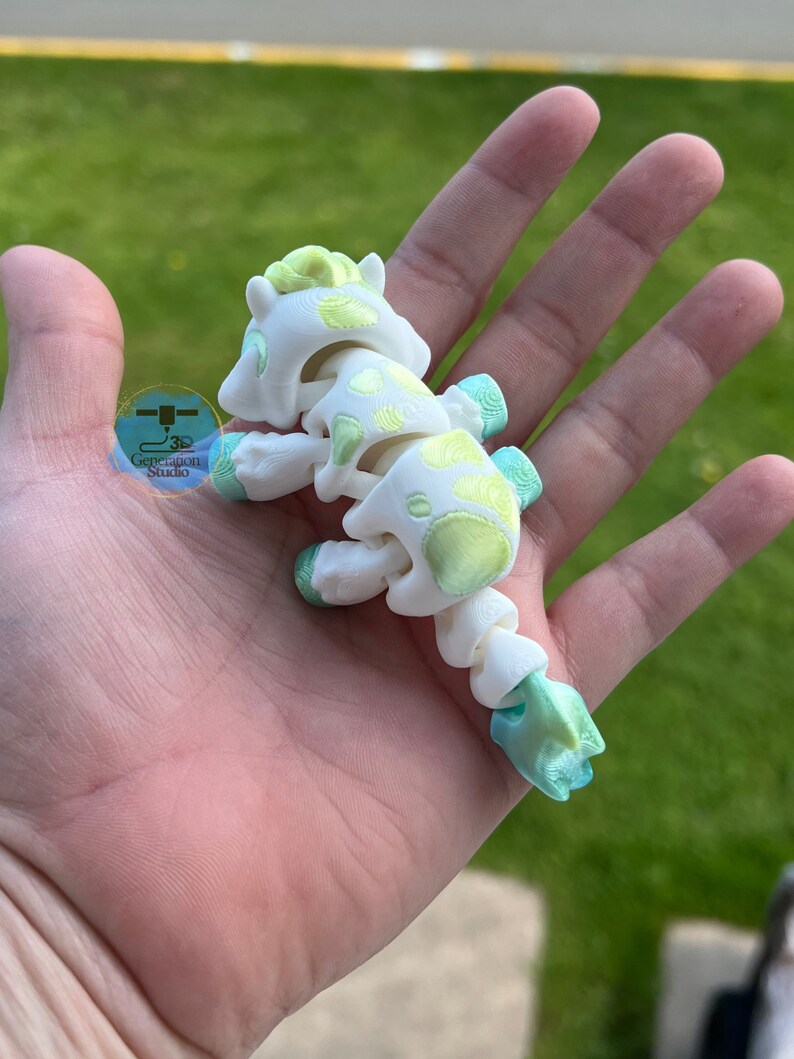 May include: A white articulated horse toy with green and yellow accents, held in a hand. The toy has a segmented body and a detailed design, with the text "Generagon Studio" visible in the background.