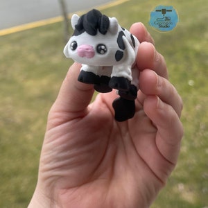 May include: A 3D-printed figurine of a cartoon cow. The cow is white with black spots, hooves, and a tuft of hair. It has large, expressive eyes and a pink muzzle. The figurine is held in a hand.