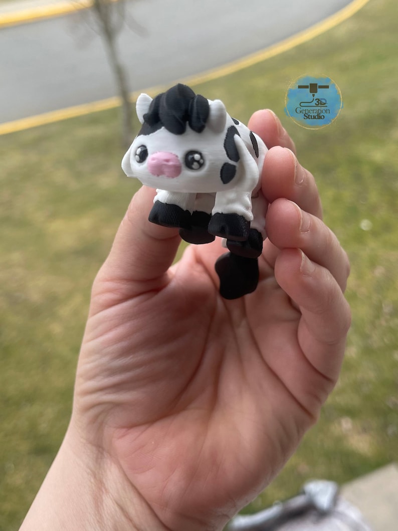 May include: A 3D-printed figurine of a cartoon cow. The cow is white with black spots, hooves, and a tuft of hair. It has large, expressive eyes and a pink muzzle. The figurine is held in a hand.