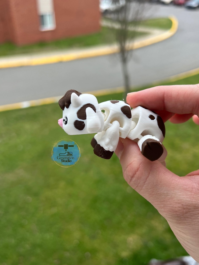 May include: A white and brown articulated cow figurine with a pink nose and dark brown spots. The cow is held in a hand against a blurred green background. The figurine has a 3D Generation Studio logo.