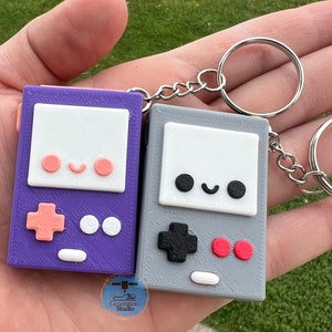 May include: Two 3D-printed keychains shaped like retro handheld game consoles. One is purple with peach accents, the other is gray with red accents. Both have a smiling face design and are attached to silver keyrings.