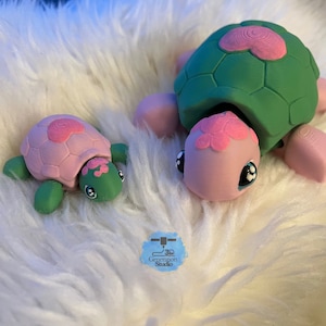 May include: Two 3D-printed turtle figurines. One is larger, the other smaller. Both have green shells with pink accents, pink bodies, and large, cartoonish eyes. The turtles are resting on a fluffy, white surface. The smaller turtle is on the left.