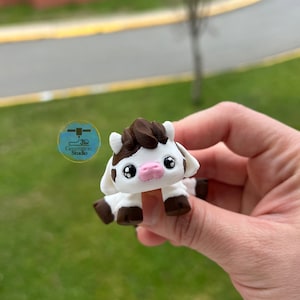 May include: A small, white and brown cow figurine with a pink nose and black eyes. The figurine is held in a hand against a blurred green background. The cow has a dark brown patch on its head and brown hooves. The logo for Generation Studio is visible.