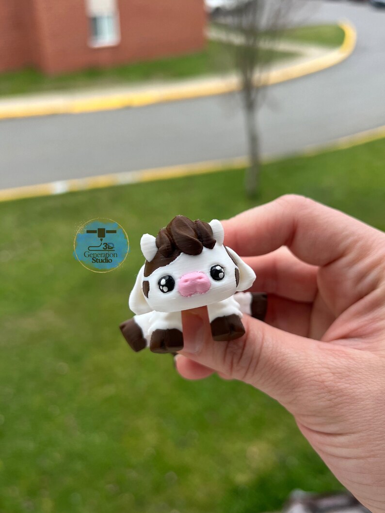 May include: A small, white and brown cow figurine with a pink nose and black eyes. The figurine is held in a hand against a blurred green background. The cow has a dark brown patch on its head and brown hooves. The logo for Generation Studio is visible.