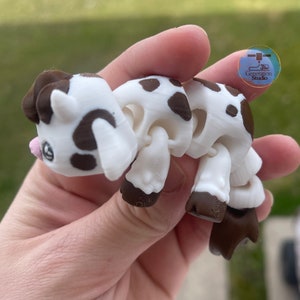 May include: A 3D-printed articulated cow toy in white and brown. The toy has a cartoonish design with brown spots, hooves, and ears. The cow is held in a hand, and the background is blurred.