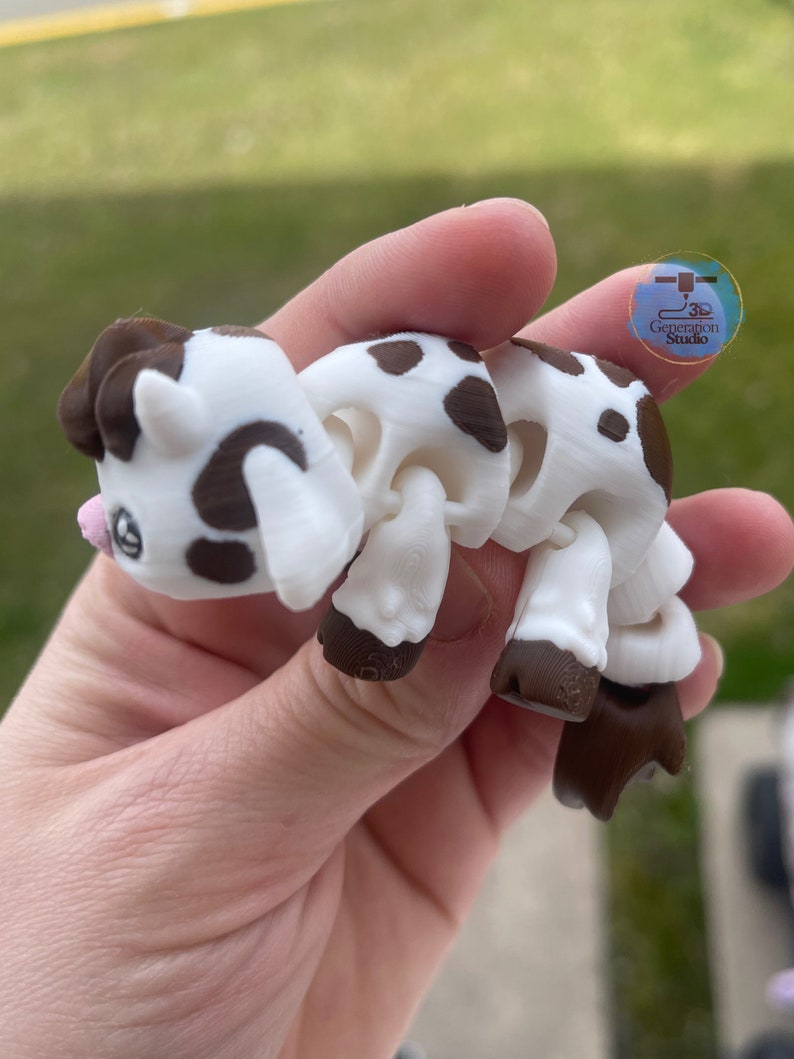 May include: A 3D-printed articulated cow toy in white and brown. The toy has a cartoonish design with brown spots, hooves, and ears. The cow is held in a hand, and the background is blurred.