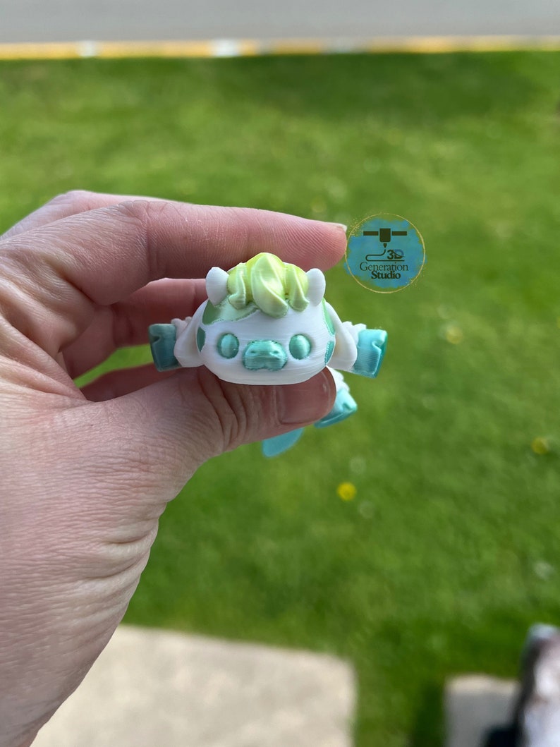 May include: A 3D-printed, white and mint-green cartoon character with a light green tuft of hair. The figure has outstretched arms and a smiling face. The logo for "3D Generation Studio" is visible in the background.