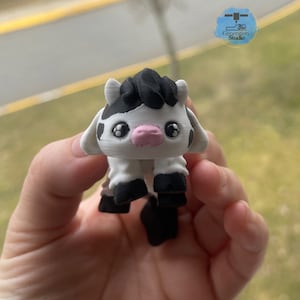 May include: A 3D-printed figurine of a cartoon cow. The cow has a white body with black spots, black hooves, and a black patch of hair on its head. It has large, expressive eyes and a pink nose. The figurine is held in a hand.