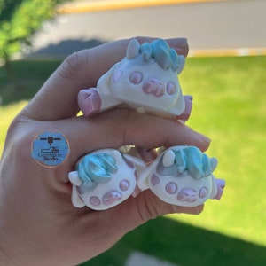 May include: Three small, white cow figurines with pink accents and light blue hair. The figurines have a cartoonish design and are held in a hand. The logo for 3D Generation Studio is visible in the image.