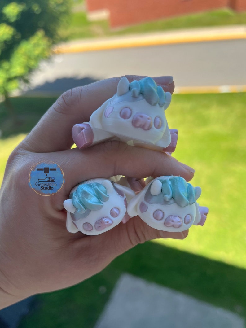 May include: Three small, white cow figurines with pink accents and light blue hair. The figurines have a cartoonish design and are held in a hand. The logo for 3D Generation Studio is visible in the image.